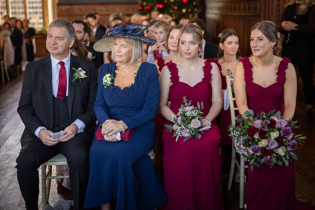 Family members and bridesmaids wearing burgundy dresses and a blue hat and dress are seated together during a wedding ceremony in a wood-panelled room.