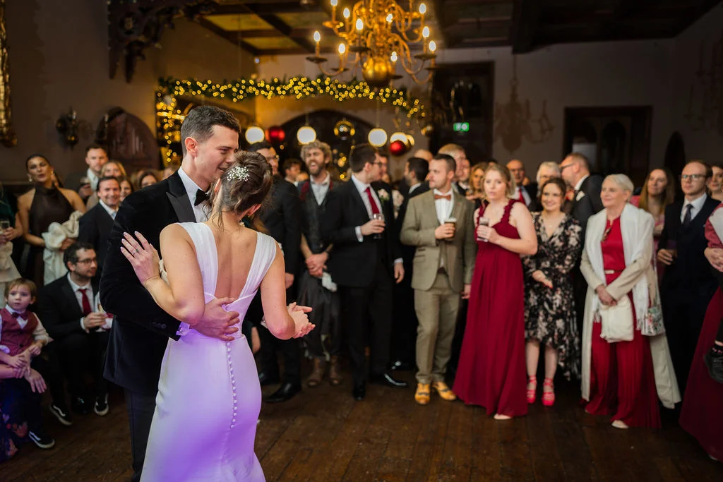 Newlyweds share their first dance surrounded by a crowd of guests in a wood-paneled ballroom illuminated by warm chandeliers and fairy lights.