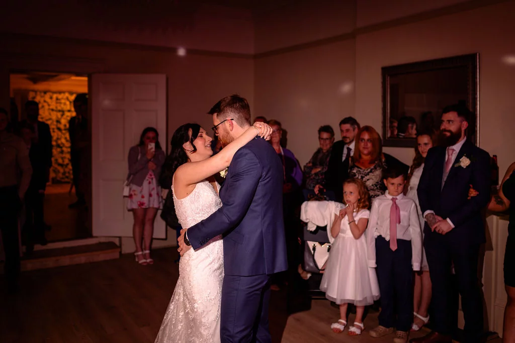 Newlyweds share their first dance surrounded by family and friends watching in a warmly lit hotel reception room.