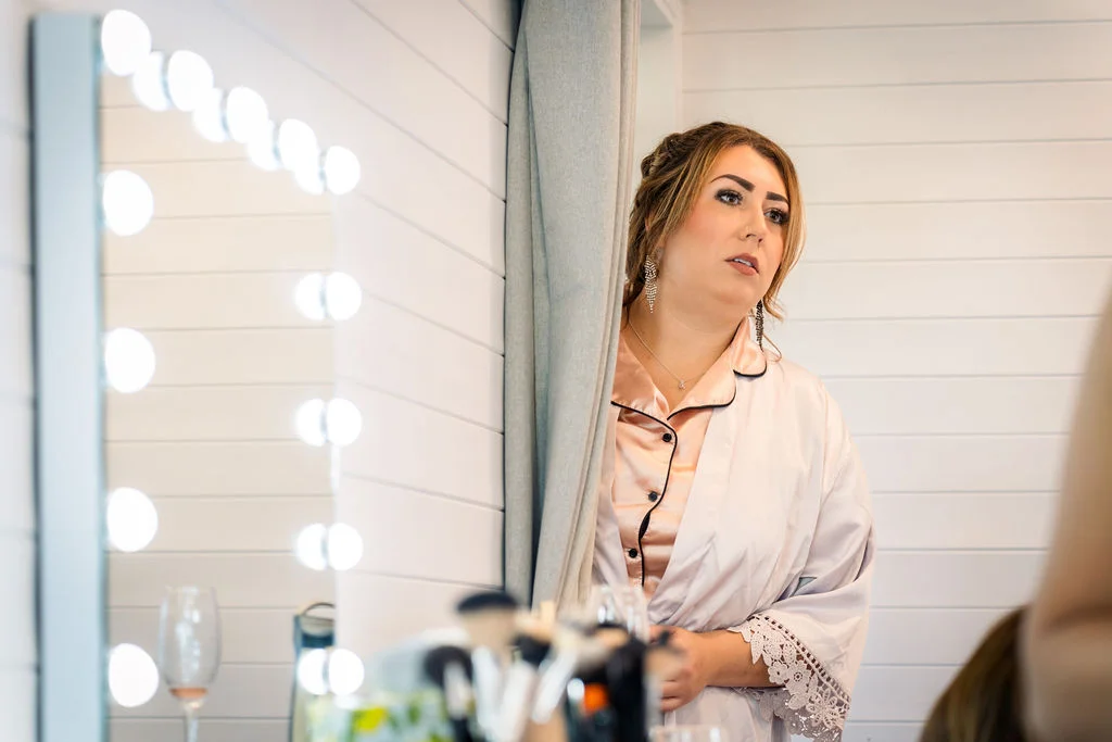 A bride in silk pyjamas and drop earrings looks into an illuminated vanity mirror while getting ready on her wedding morning.