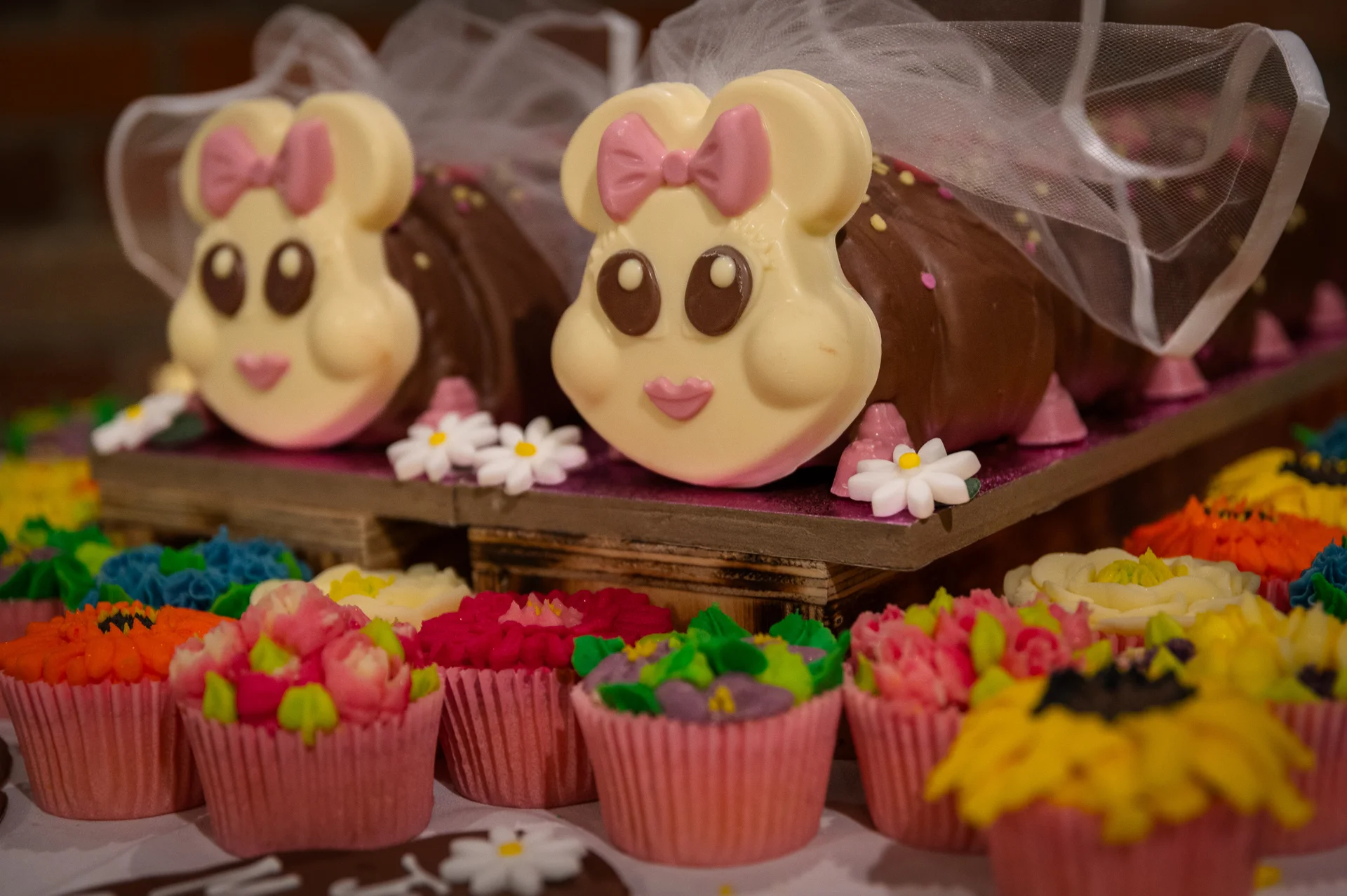 A whimsical wedding dessert display features novelty animal chocolate cakes adorned with tulle veils, surrounded by colourful floral cupcakes.