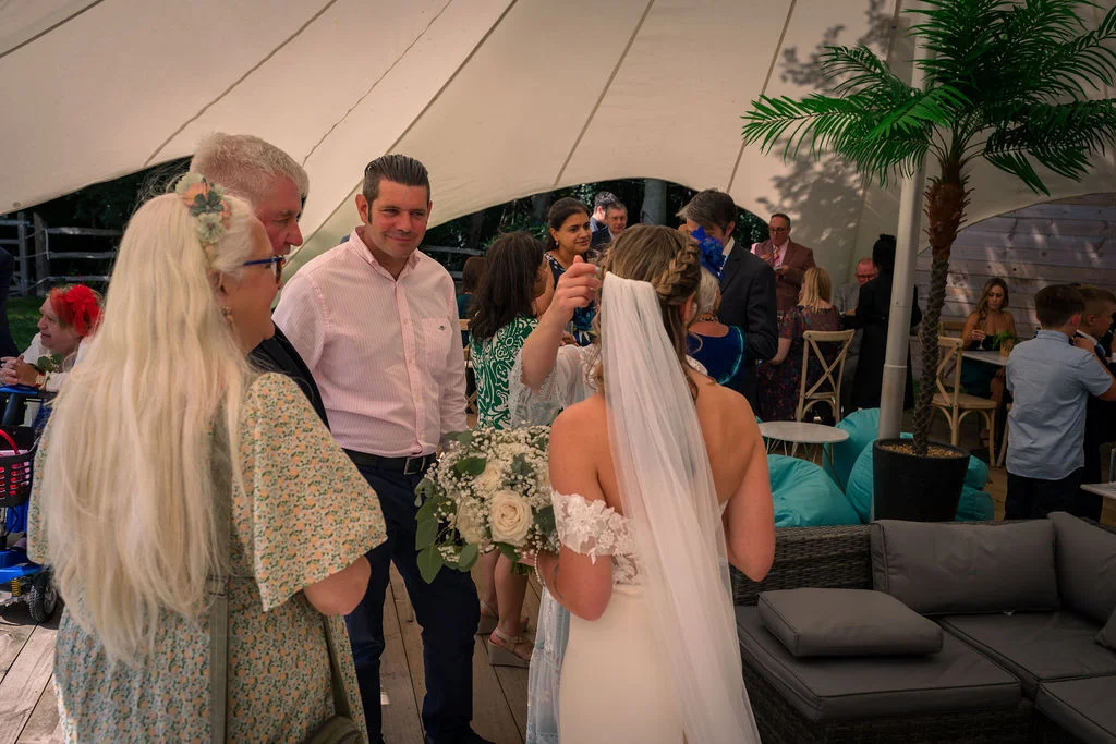 A bride in a white off-the-shoulder gown holding a bouquet chats with family and guests mingling inside a white stretch tent marquee.