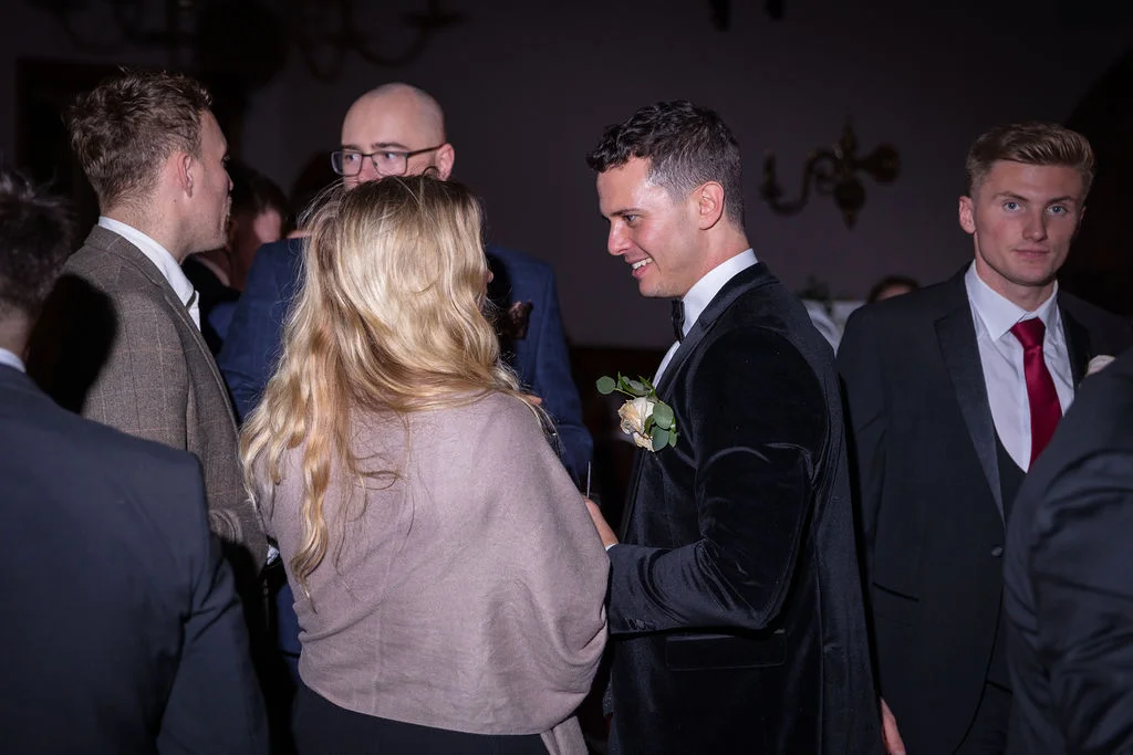 A groom in a velvet tuxedo smiling and chatting with guests during an evening wedding reception.
