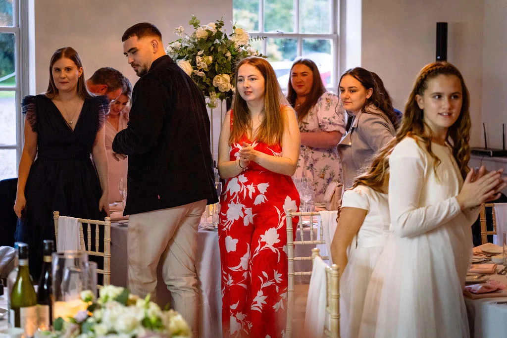 Wedding guests stand and applaud during speeches at a wedding reception featuring white and green floral centrepieces.