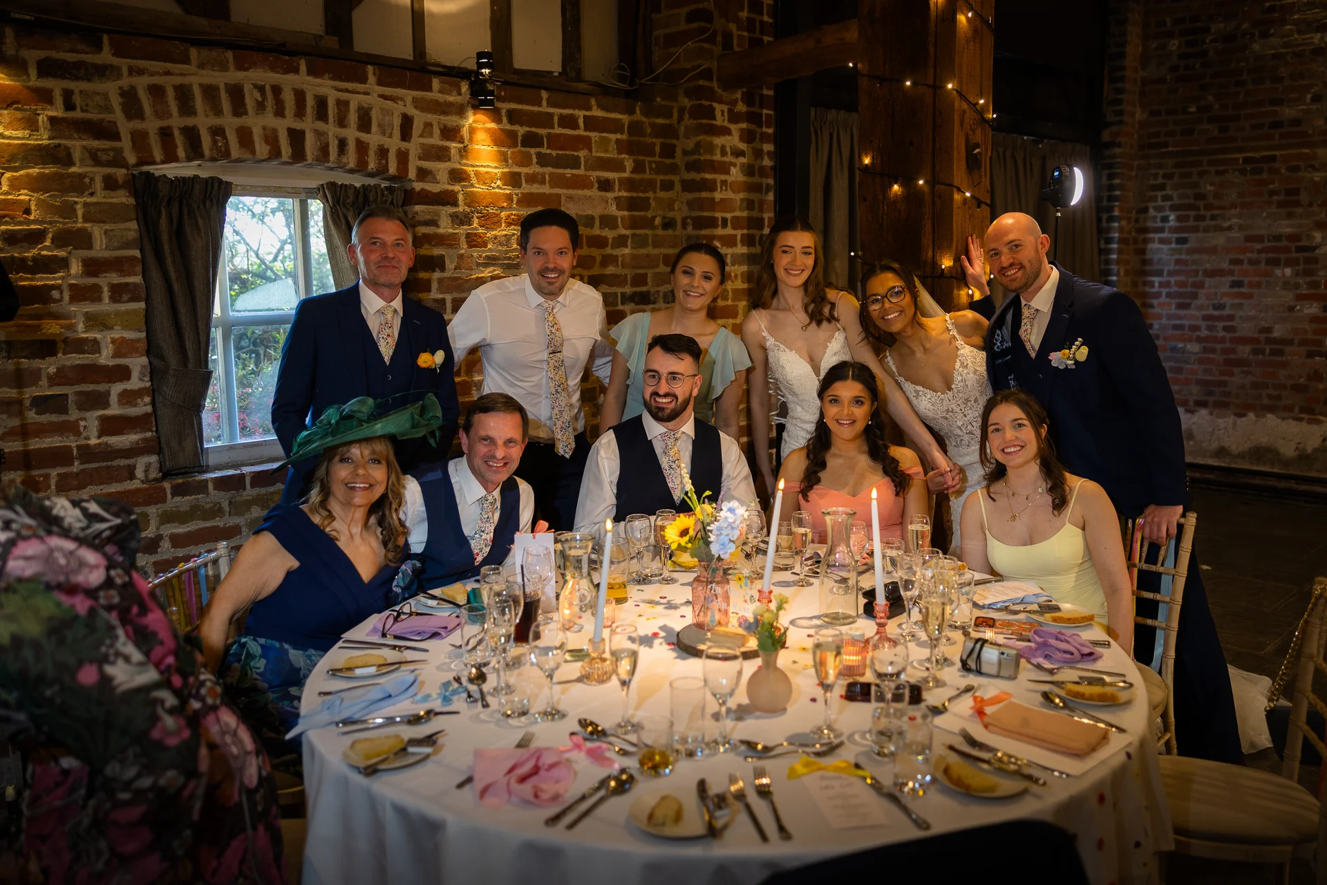 A smiling wedding party and guests gather around a warmly lit round table inside a rustic brick barn venue.