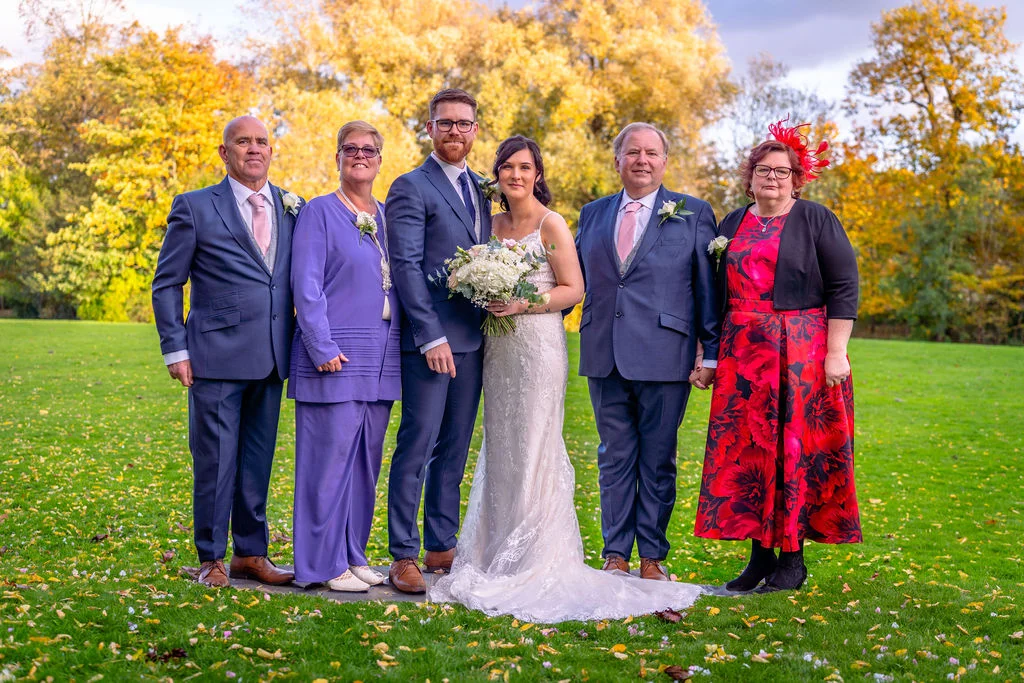 A newlywed couple posing outdoors on a lush green lawn with family members surrounded by vibrant autumn foliage.