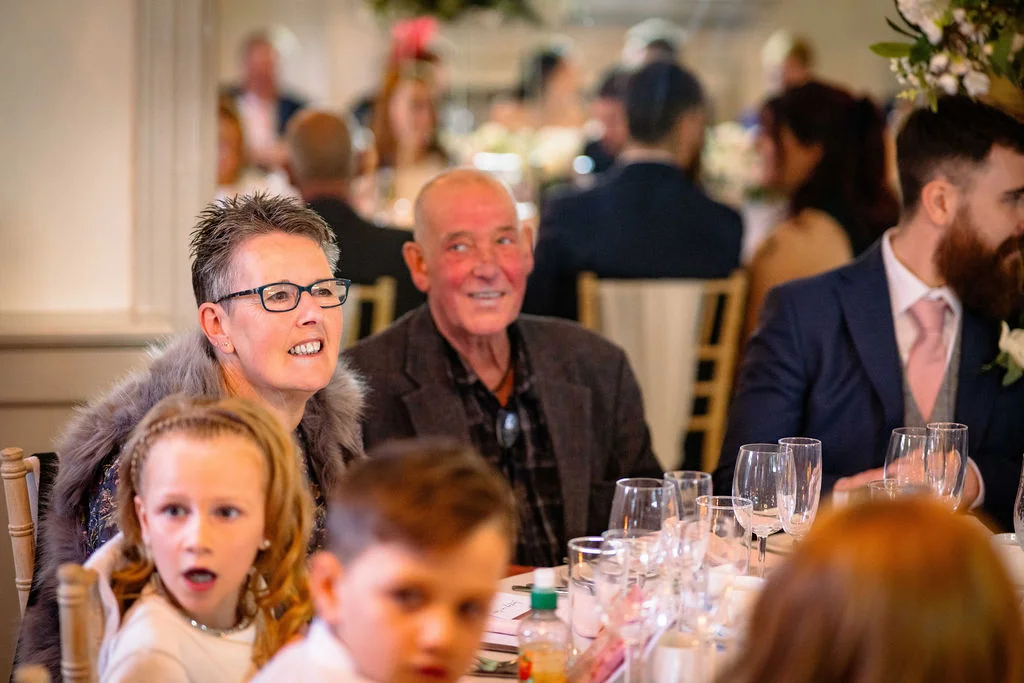Wedding guests including smiling older adults and children enjoying speeches at a beautifully set reception table.