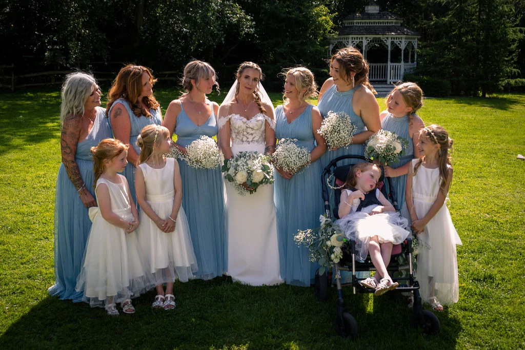 A smiling bride stands centre in an a-line lace gown holding a bouquet, surrounded by her bridesmaids in dusty blue dresses and flower girls in white on a sunlit lawn.