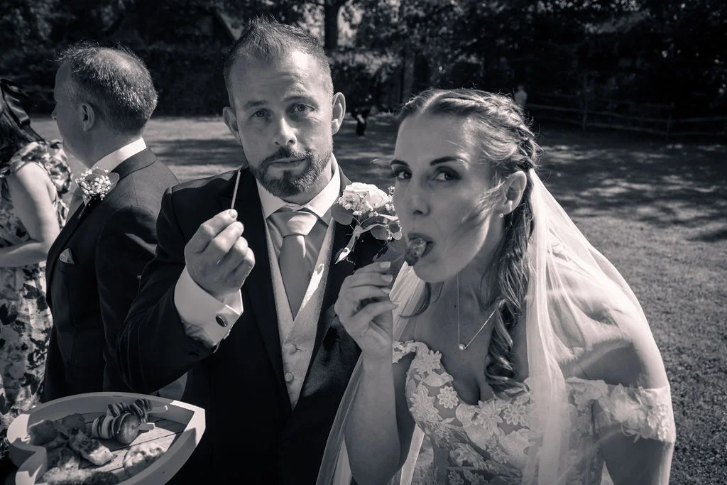 A bride and groom share a playful moment eating canapes outdoors on a sunny wedding day, captured in a striking black and white edit.