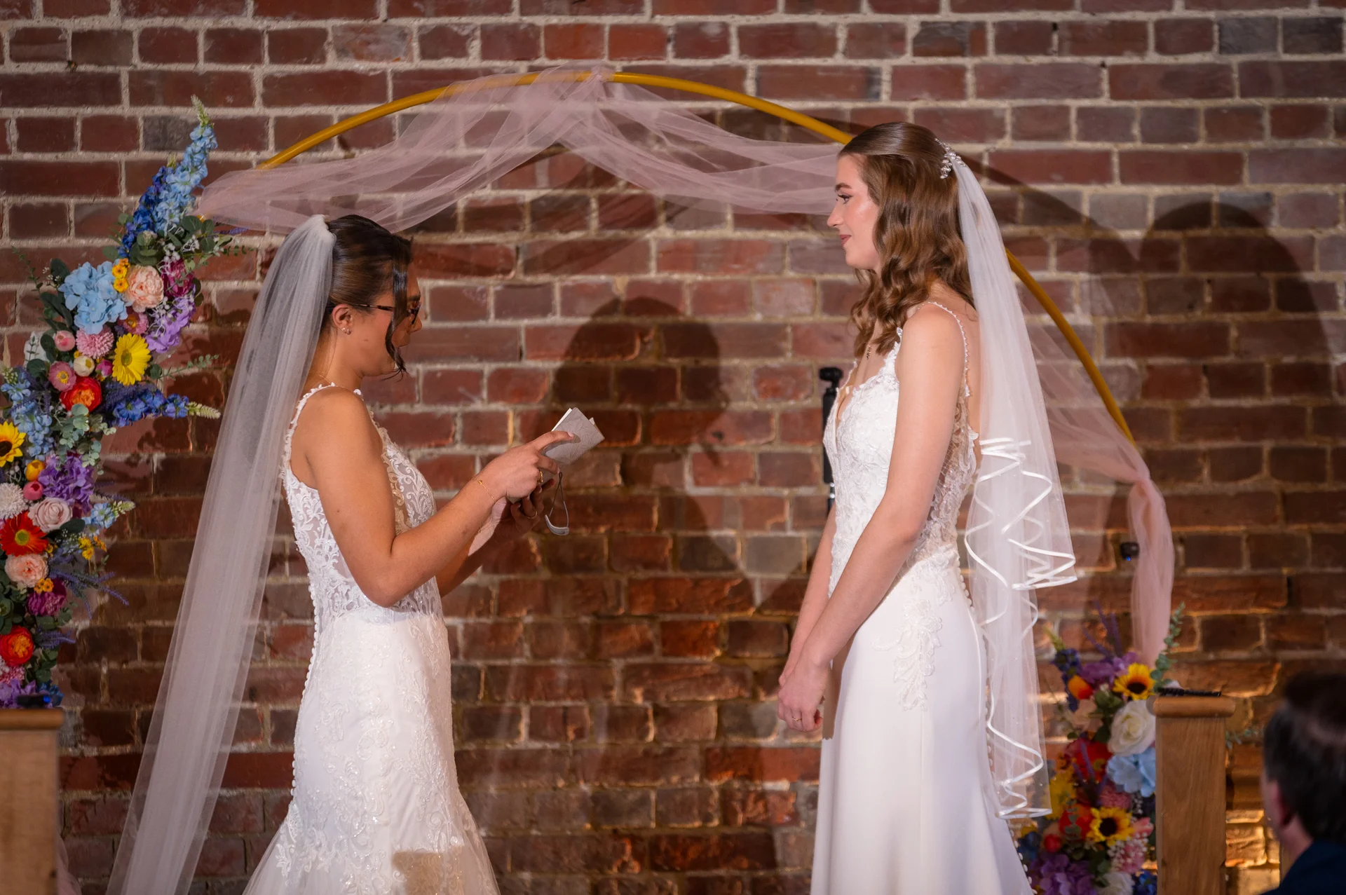Two brides stand together reading their vows during a ceremony framed by an arch and exposed brick wall.