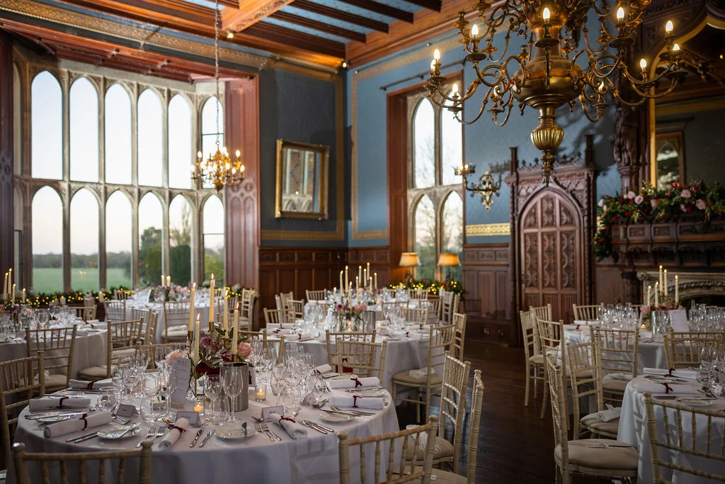 An elegant wedding breakfast reception setup inside a grand stately home ballroom with tall arched windows, wooden panelling, and beautifully laid round tables lit by warm candlelight and chandeliers.
