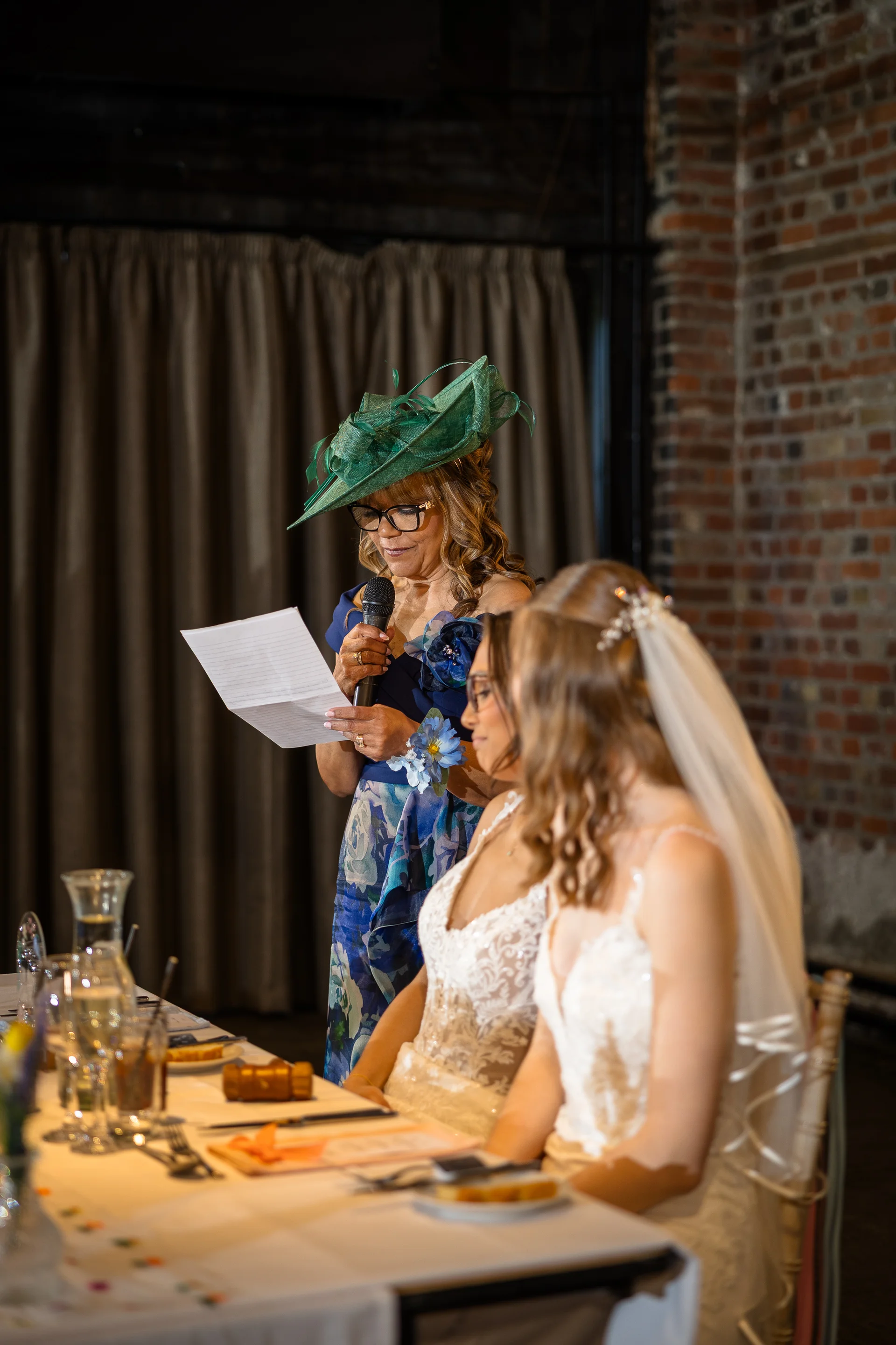 An older woman wearing a green fascinator delivers a heartfelt speech from notes at the top table during a wedding reception, while two brides listen attentively beside her.