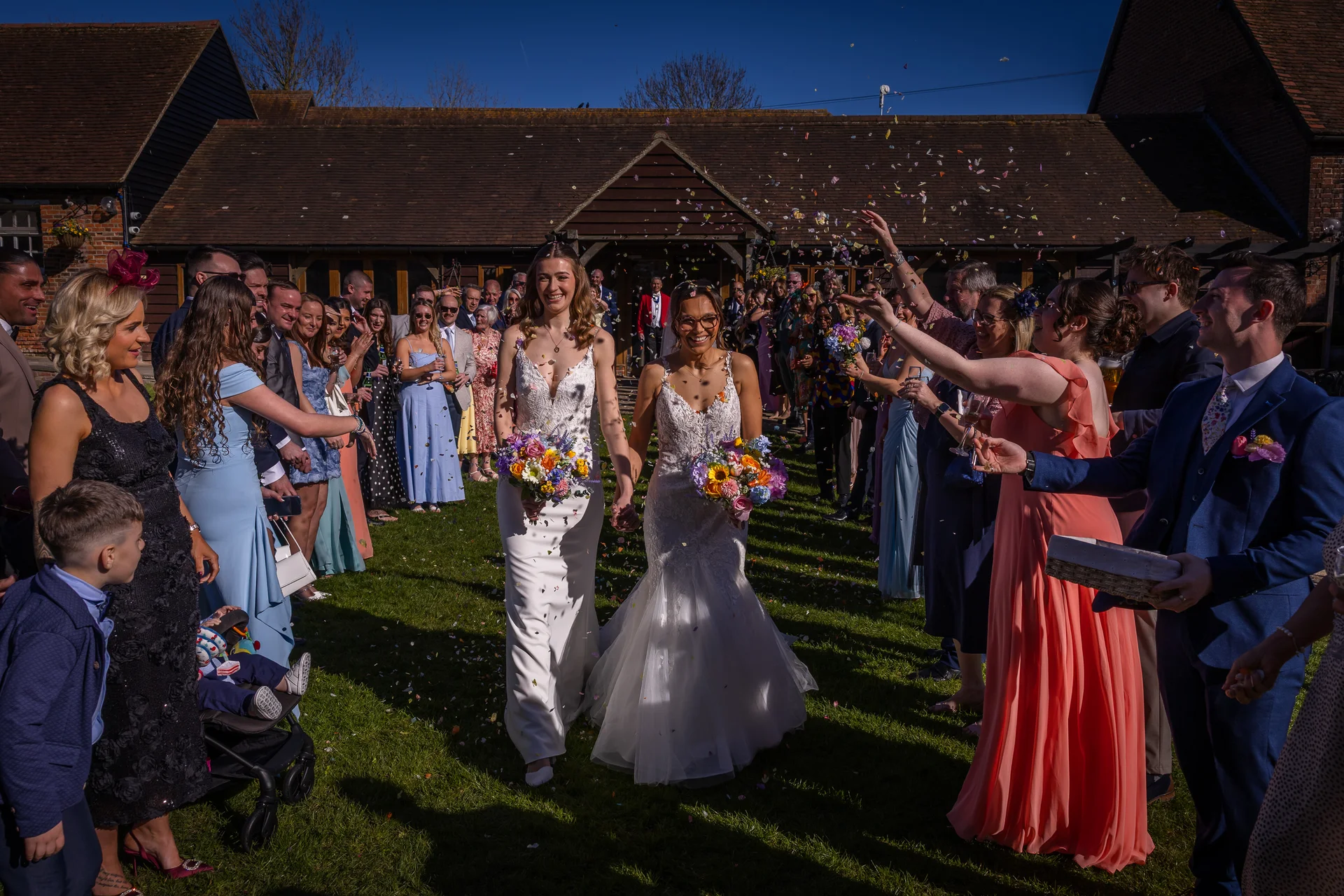Two smiling brides holding colorful bouquets walk hand in hand through a cheering crowd of wedding guests tossing confetti outside a rustic barn venue under a bright blue sky.
