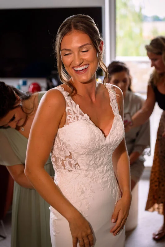 A joyful bride laughs happily as her bridesmaid fastens her lace-detailed wedding gown during morning bridal preparations.