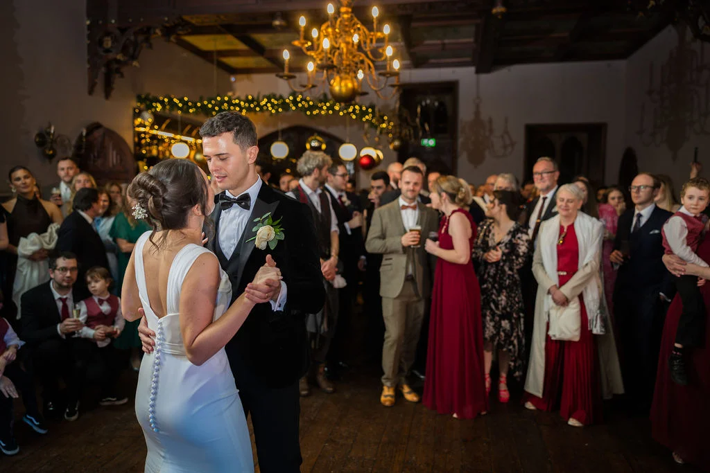 Newlyweds share their romantic first dance surrounded by family and friends in a grand hall illuminated by an ornate chandelier and warm fairy lights.