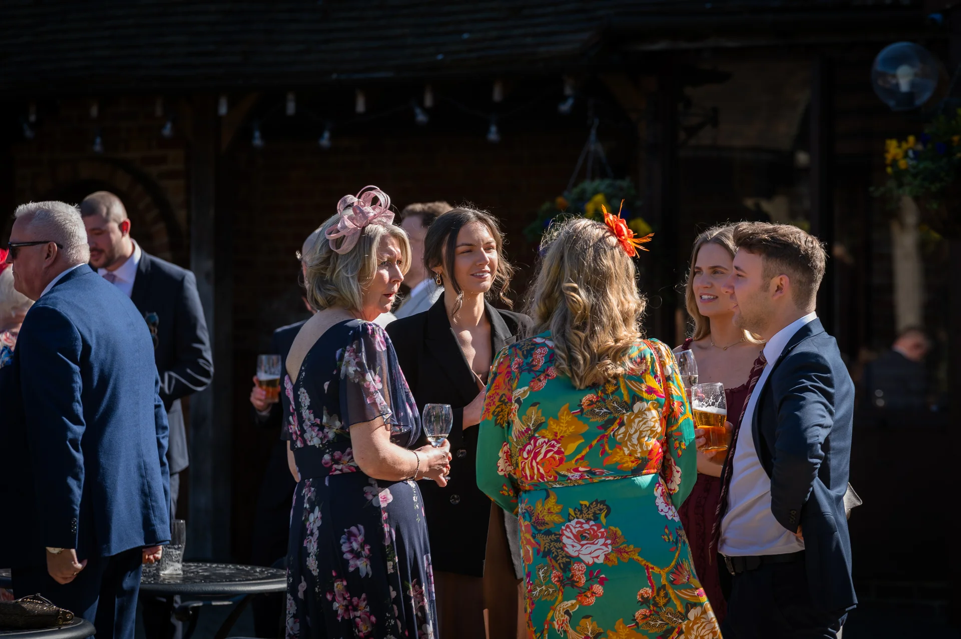 Wedding guests mingle and chat outdoors in the warm sunshine during the drinks reception outside a rustic venue.