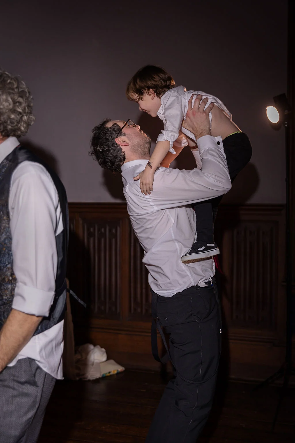A joyful guest lifts a young child up into the air during the wedding reception celebration in a panelled room.