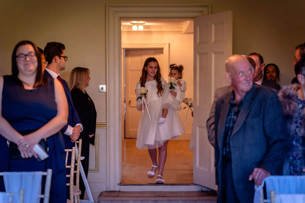 A young flower girl carrying another toddler walks through an open doorway as wedding guests look on expectantly.