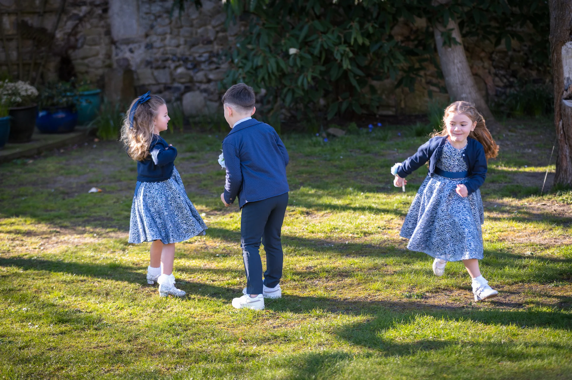 Young flower girls and a page boy in matching blue outfits play joyfully on a sunlit lawn during a wedding reception.
