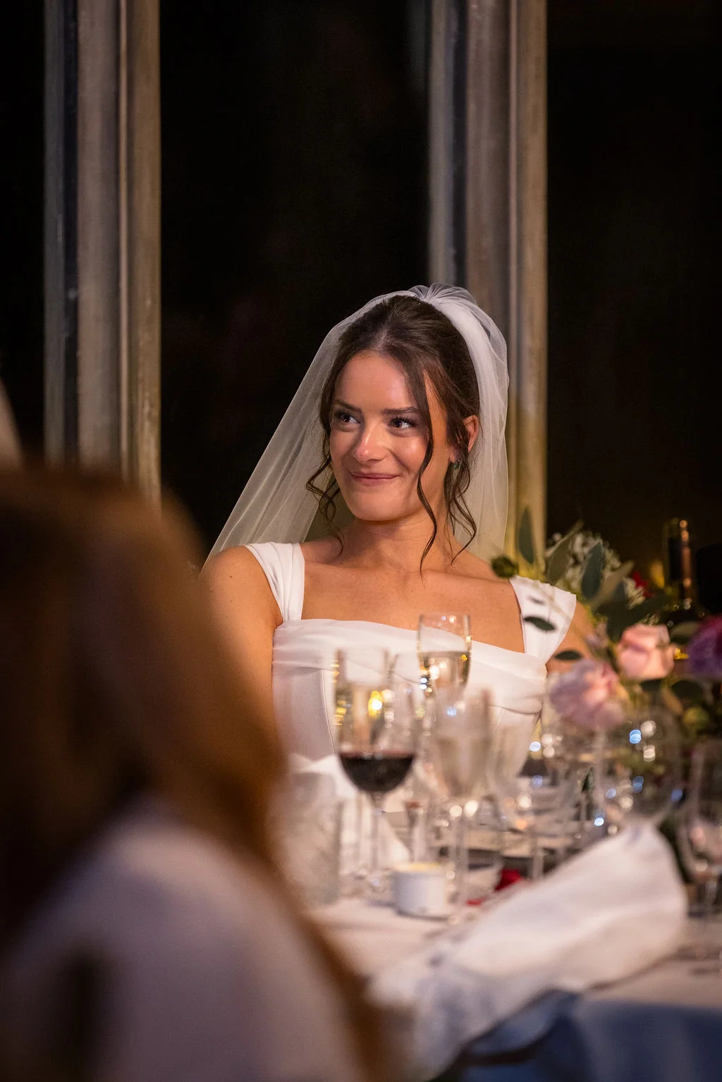 A smiling bride wearing a square neckline dress and a veil sits at the wedding breakfast table during the speeches.