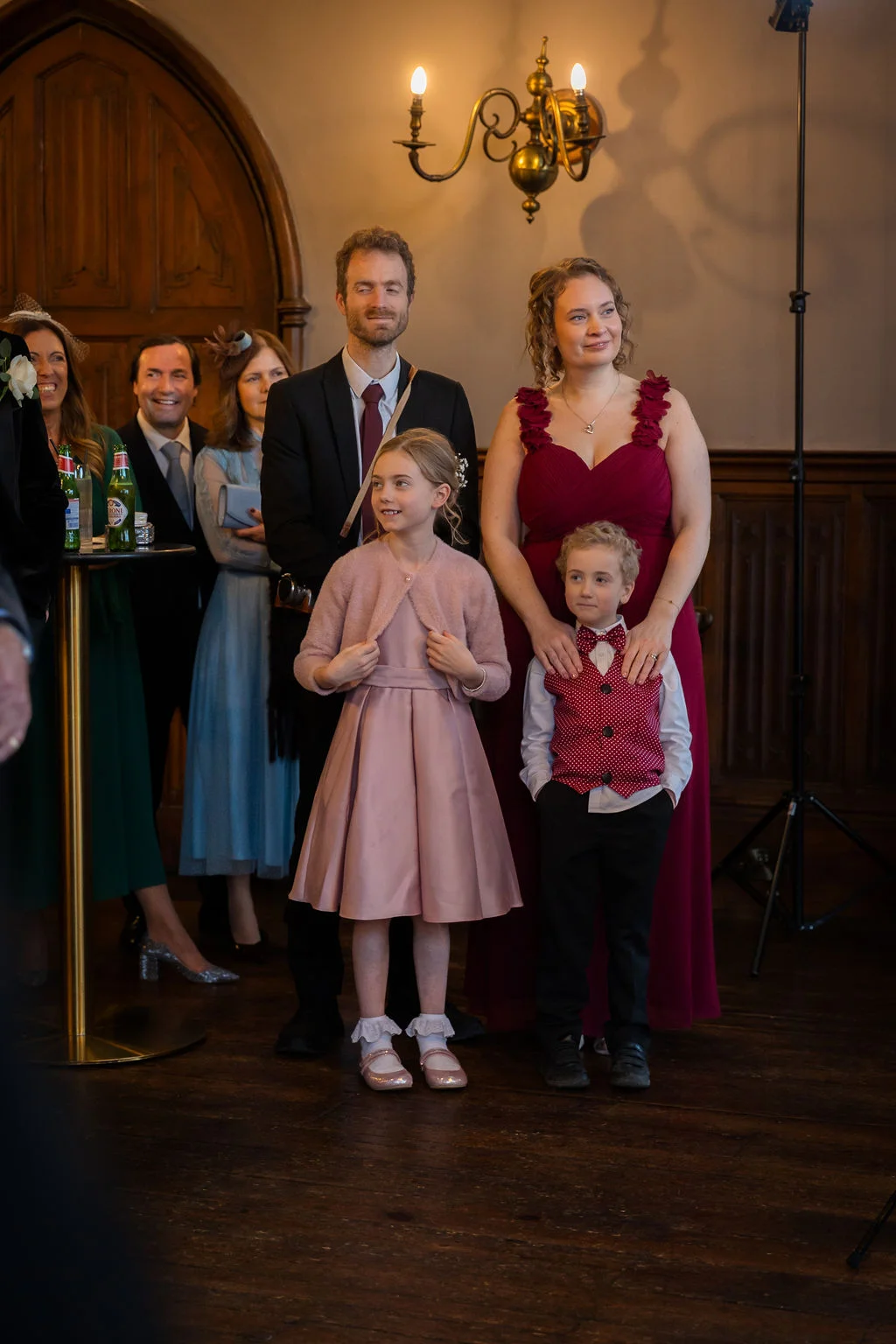 A group of wedding guests and family members, including smiling children in matching pink and red attire, stand together watching speeches in a wood-panelled room.