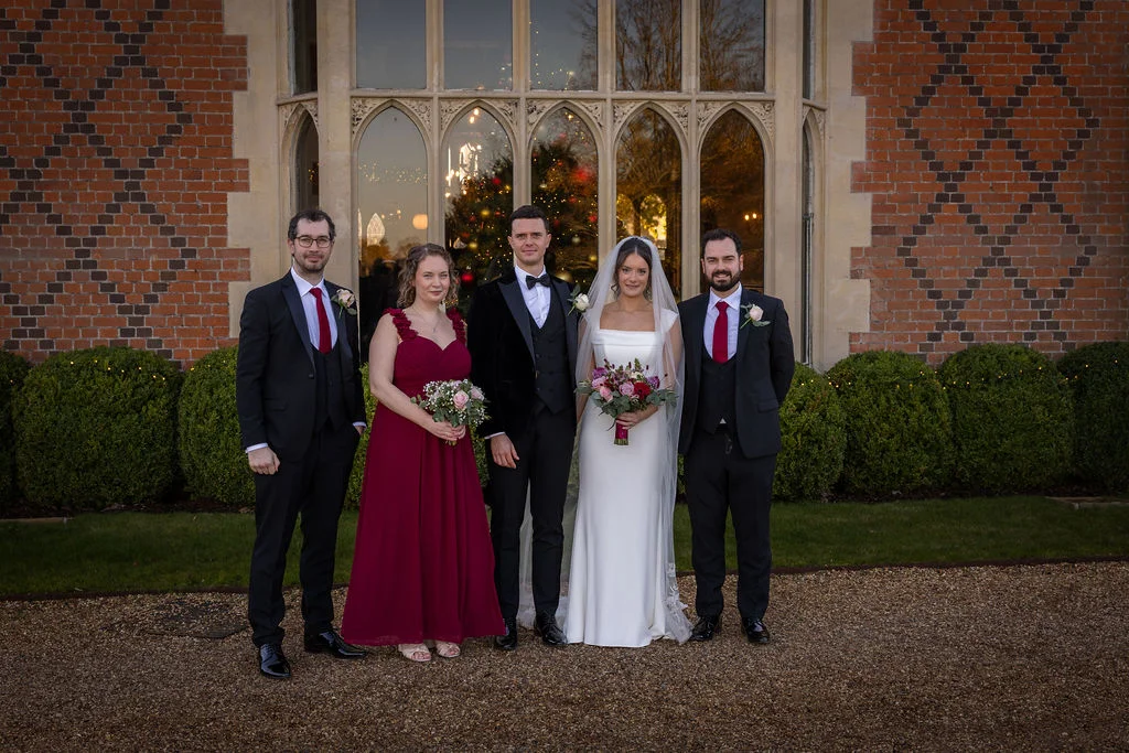 A newlywed couple posing with their wedding party outside a grand red brick manor house with large arched windows.