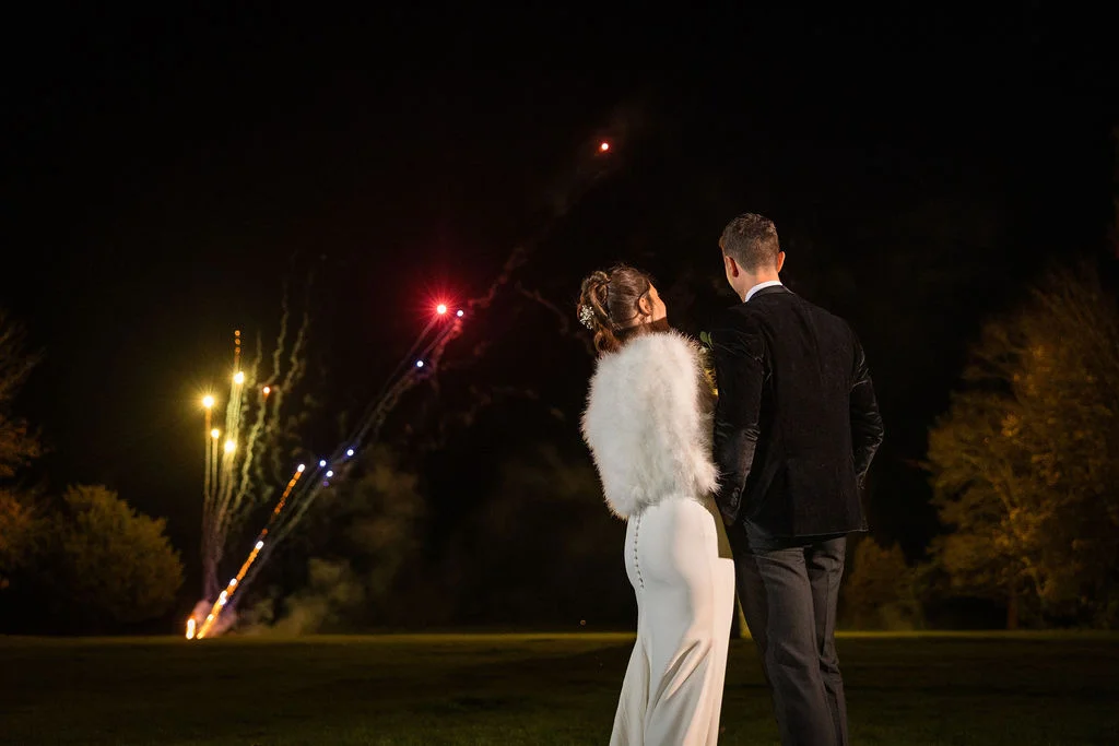 A newlywed couple stands outdoors at night, admiring a vibrant fireworks display lighting up the dark sky.
