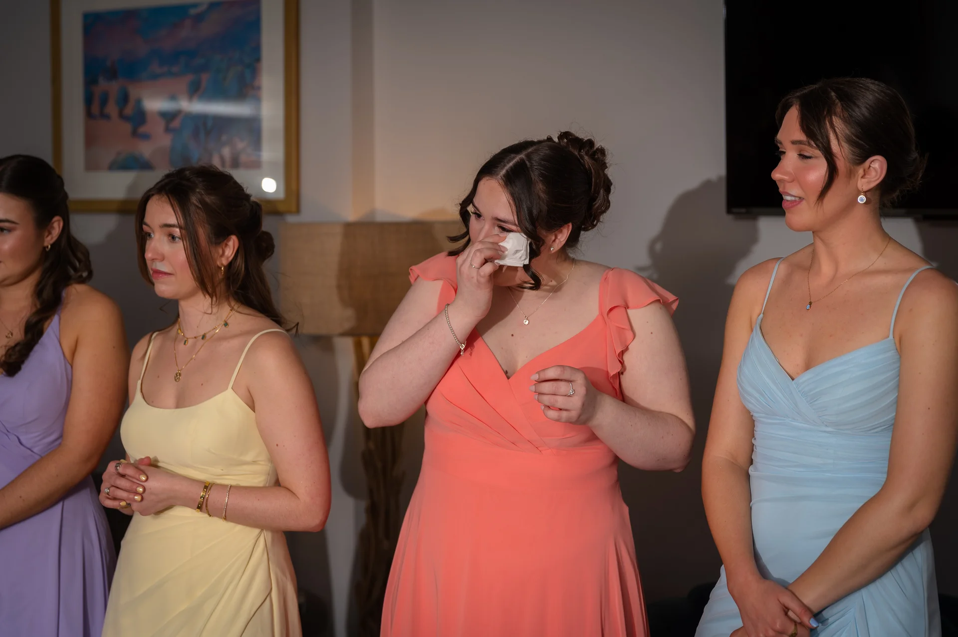 An emotional bridesmaid wipes away happy tears with a tissue while standing alongside her fellow bridesmaids wearing pastel-coloured dresses during wedding speeches.