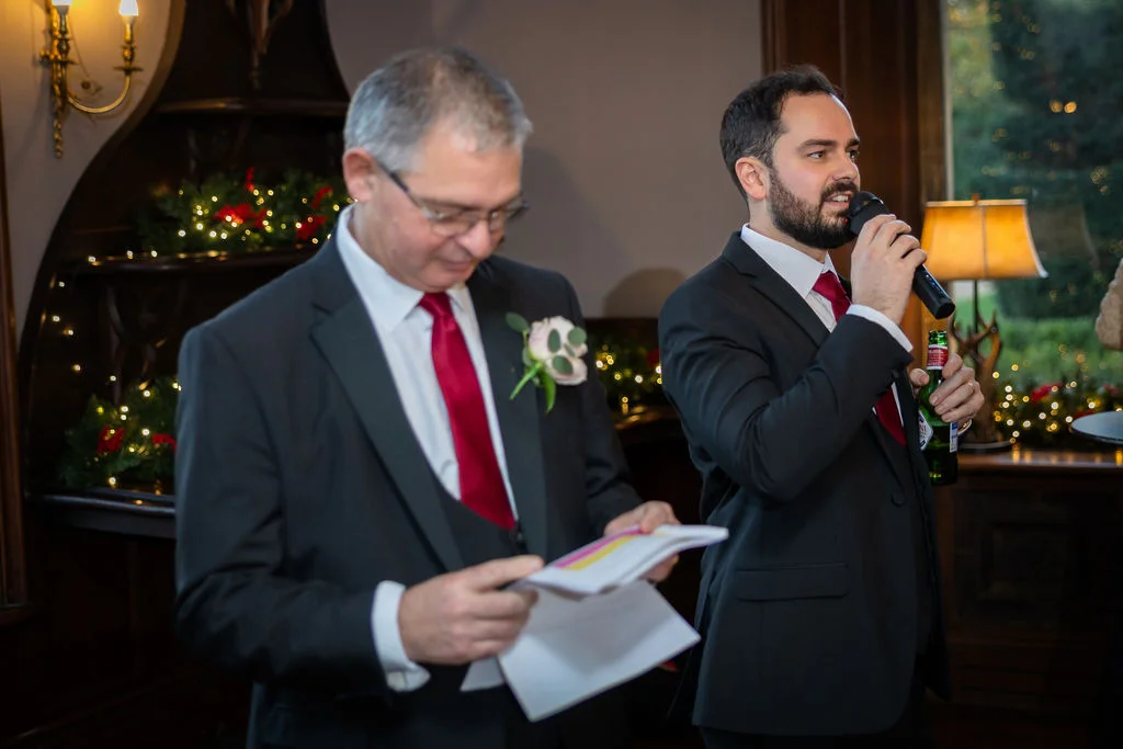 Two men in dark suits with red ties deliver wedding speeches indoors, with one holding speech notes and the other speaking into a microphone.