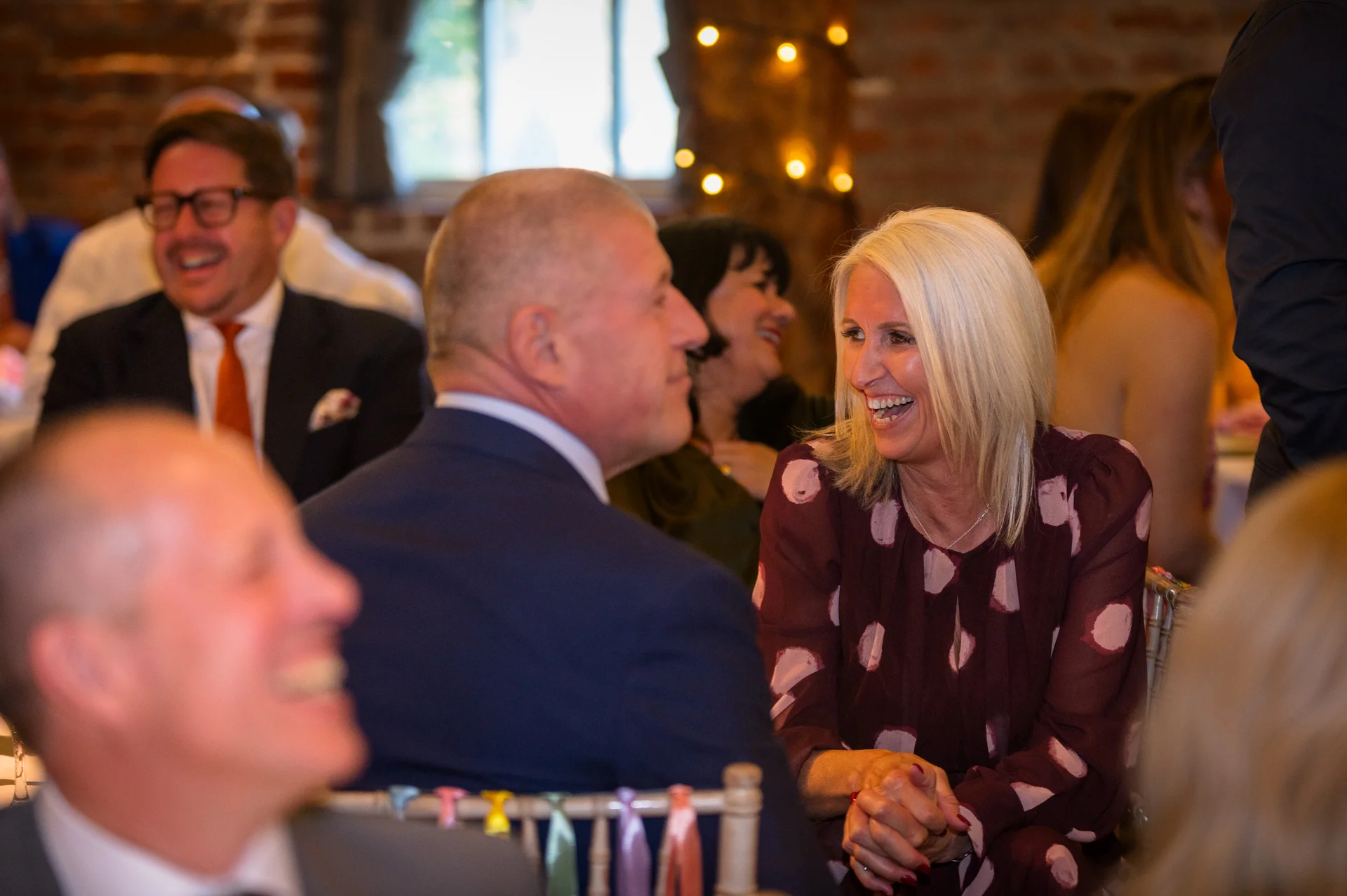 A guest at a wedding reception laughs joyfully during the speeches in a rustic barn venue with exposed brick walls.