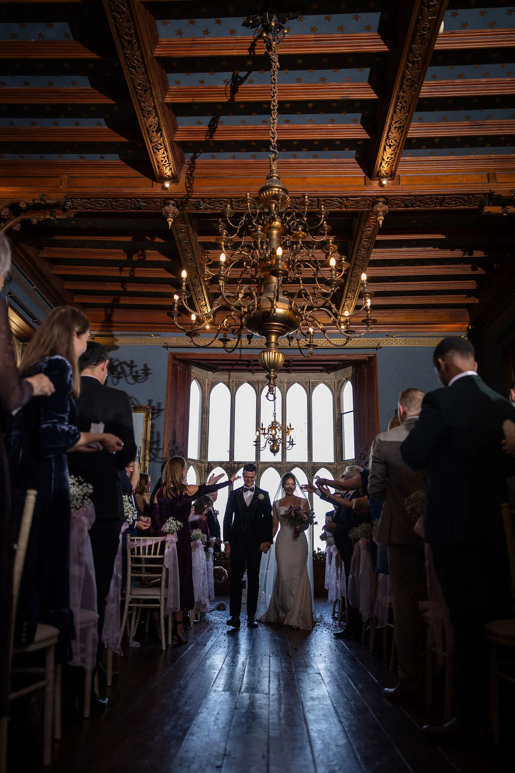 Newlyweds walk down the aisle together as smiling guests shower them with confetti in a grand historic hall featuring a striking wooden ceiling and large arched windows.