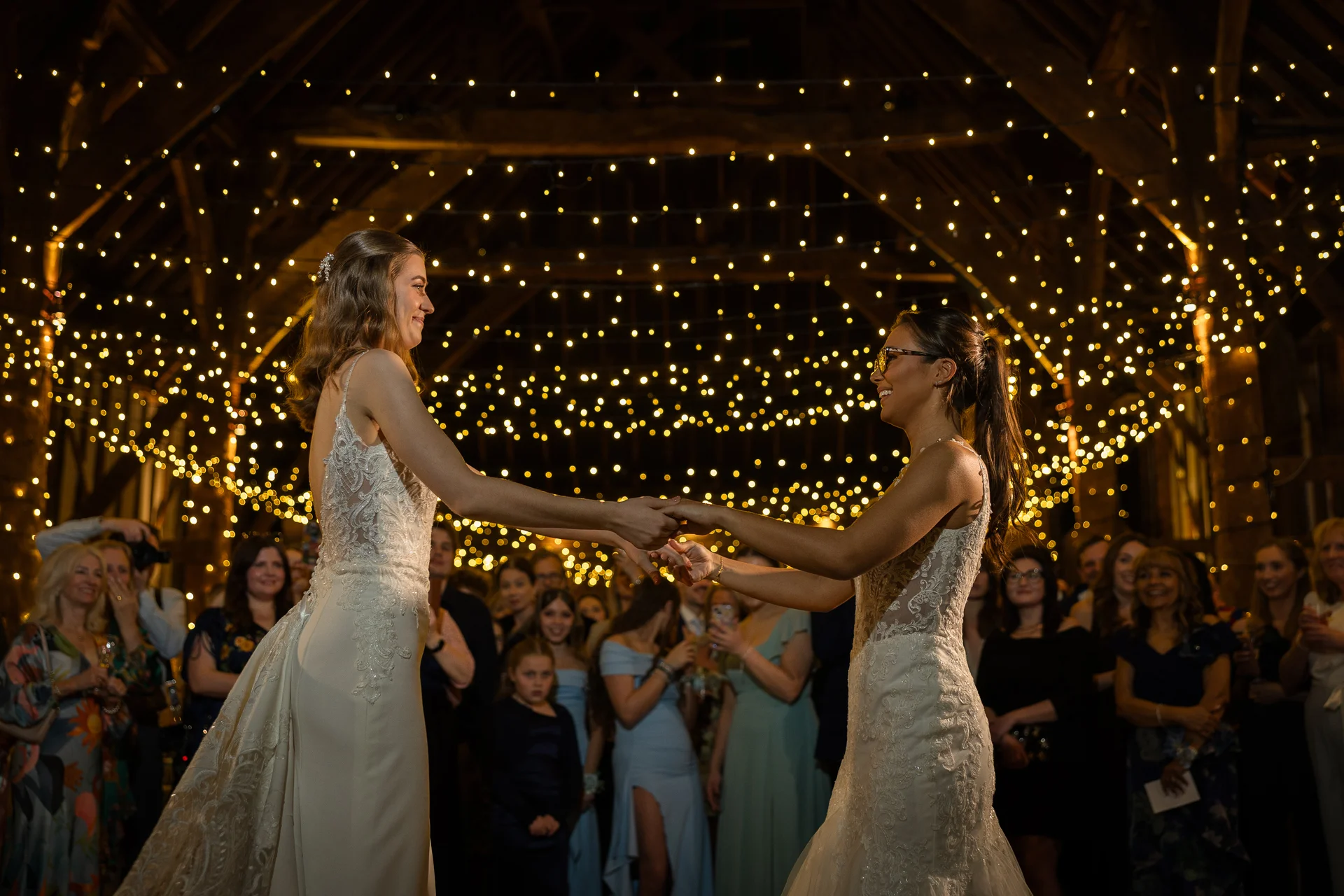 Two brides share their first dance holding hands beneath a glowing canopy of fairy lights inside a rustic oak framed barn.