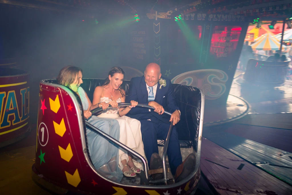 A laughing bride and groom enjoy a ride on a fairground attraction surrounded by colourful lights and smoke.