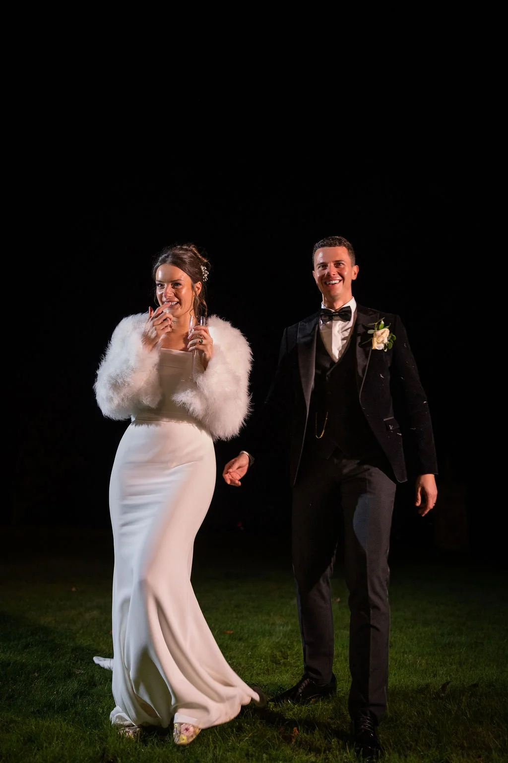 A newlywed couple smiling and walking together outdoors at night, with the bride wearing a white crepe gown and a faux fur stole while holding a glass of champagne, and the groom in a classic black tuxedo.