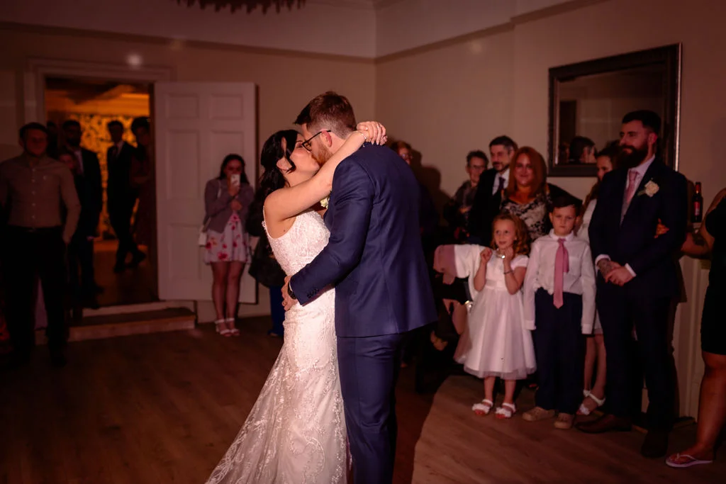Newlyweds share an intimate first kiss during their evening reception, surrounded by watching guests inside a warmly lit venue.