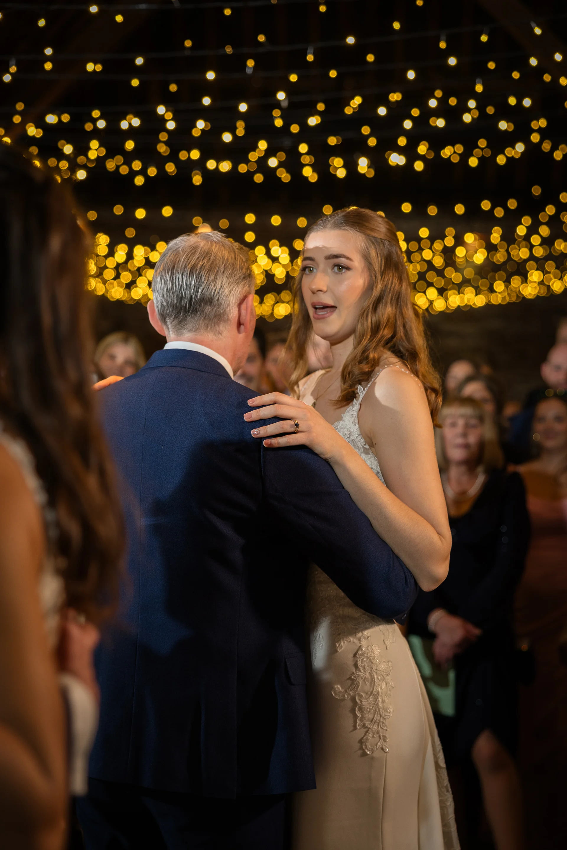 A bride shares a tender father-daughter dance under a magical canopy of glowing fairy lights inside a rustic barn venue.