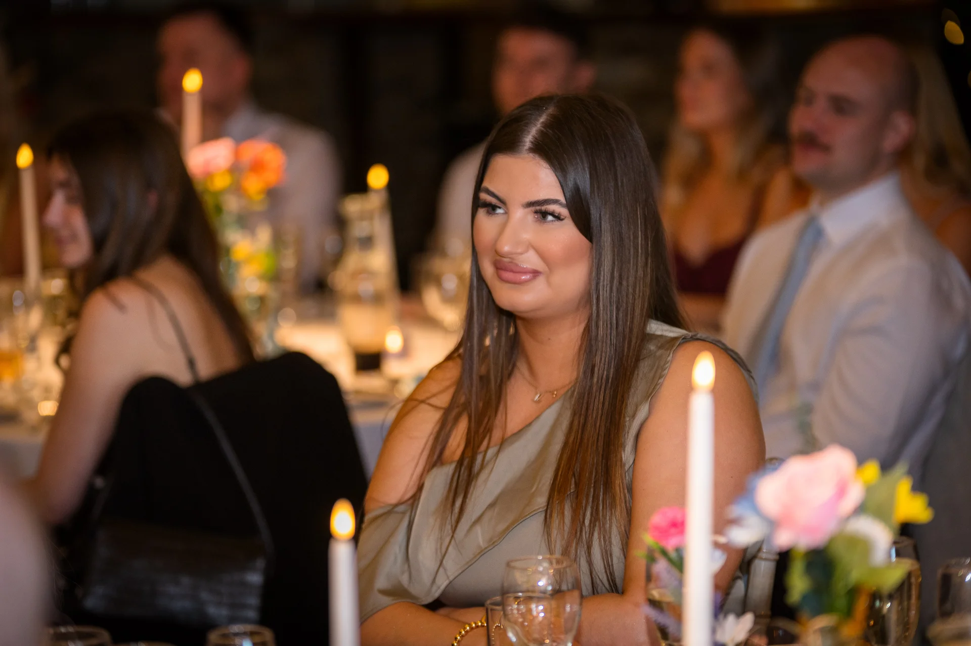 A wedding guest with long dark hair smiling during the wedding breakfast in a warm candlelit reception room.