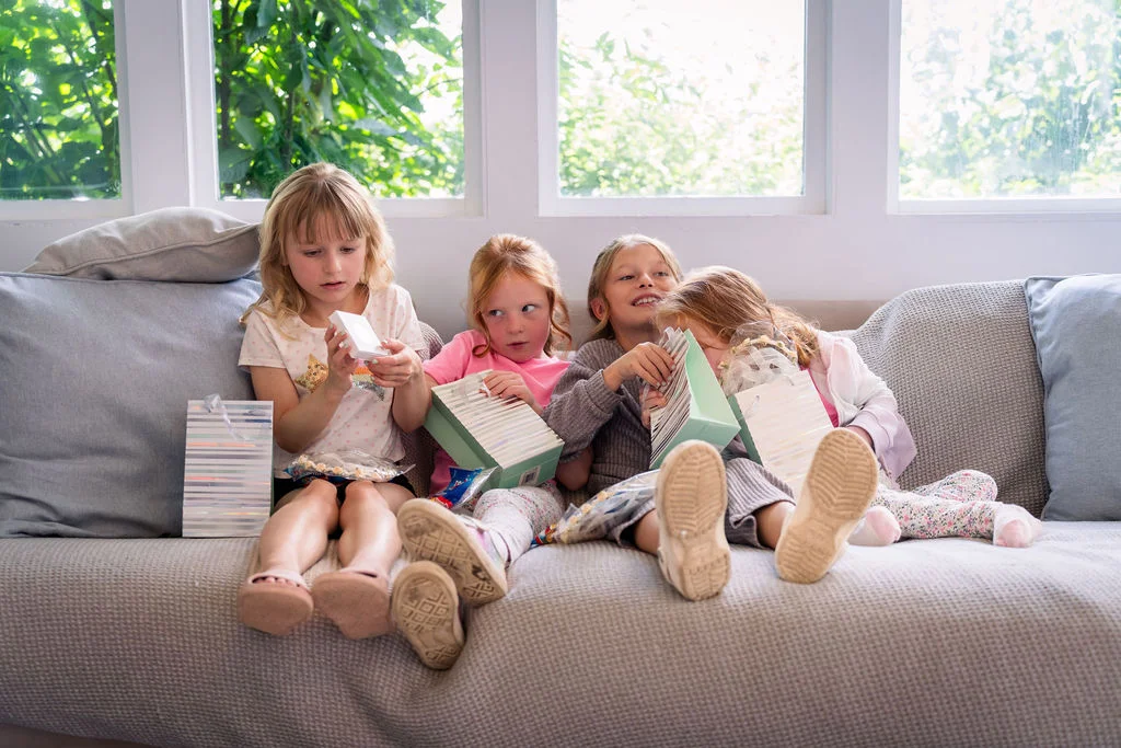 Four young flower girls sit on a grey sofa near a large window, happily opening gift bags and examining small presents.