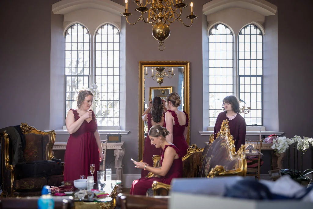 Bridesmaids in burgundy dresses gather in an elegant historic room with tall arched windows and ornate gold mirrors during bridal preparations.