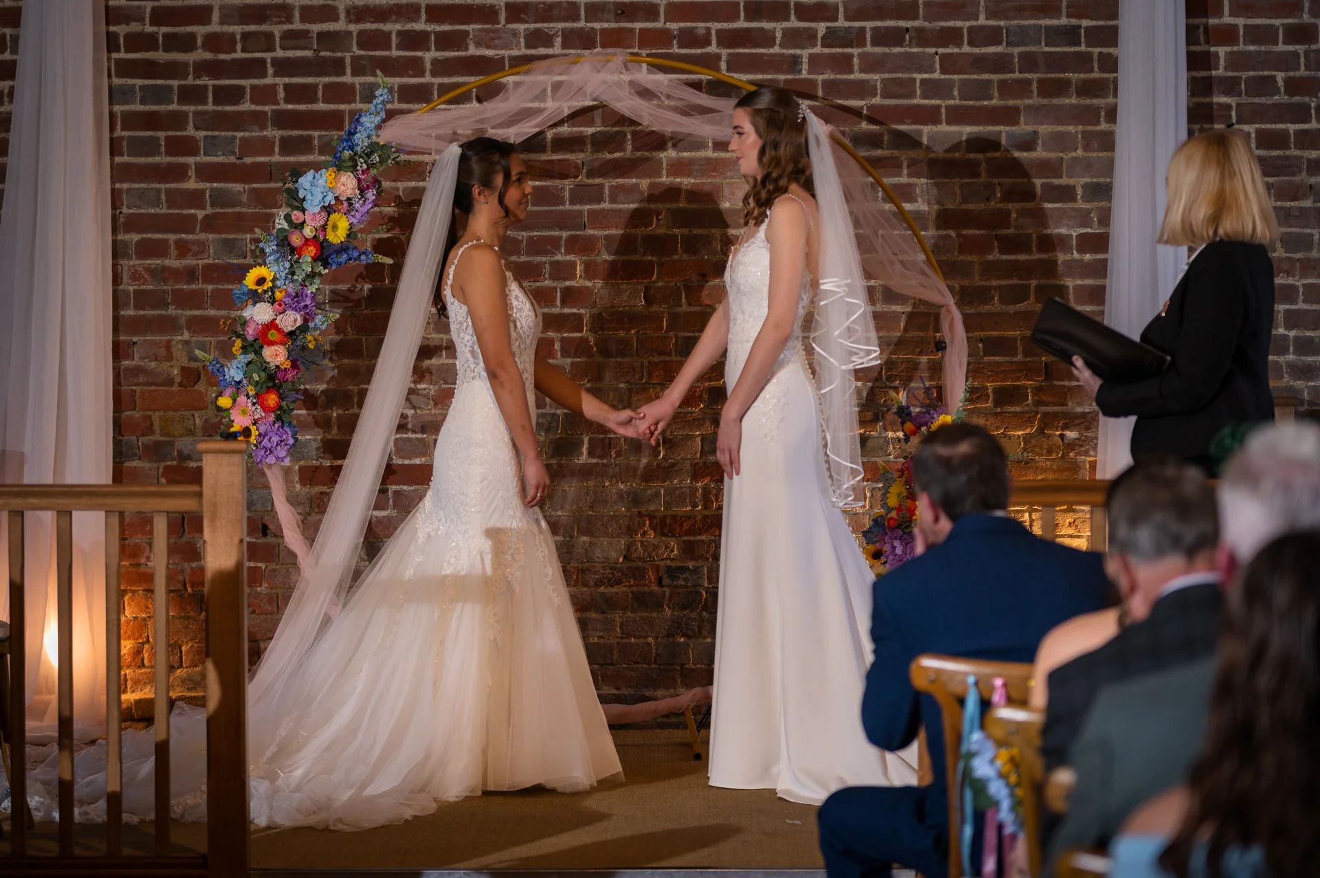 Two brides in white wedding dresses hold hands during an indoor ceremony in front of an exposed brick wall decorated with a floral hoop arch.