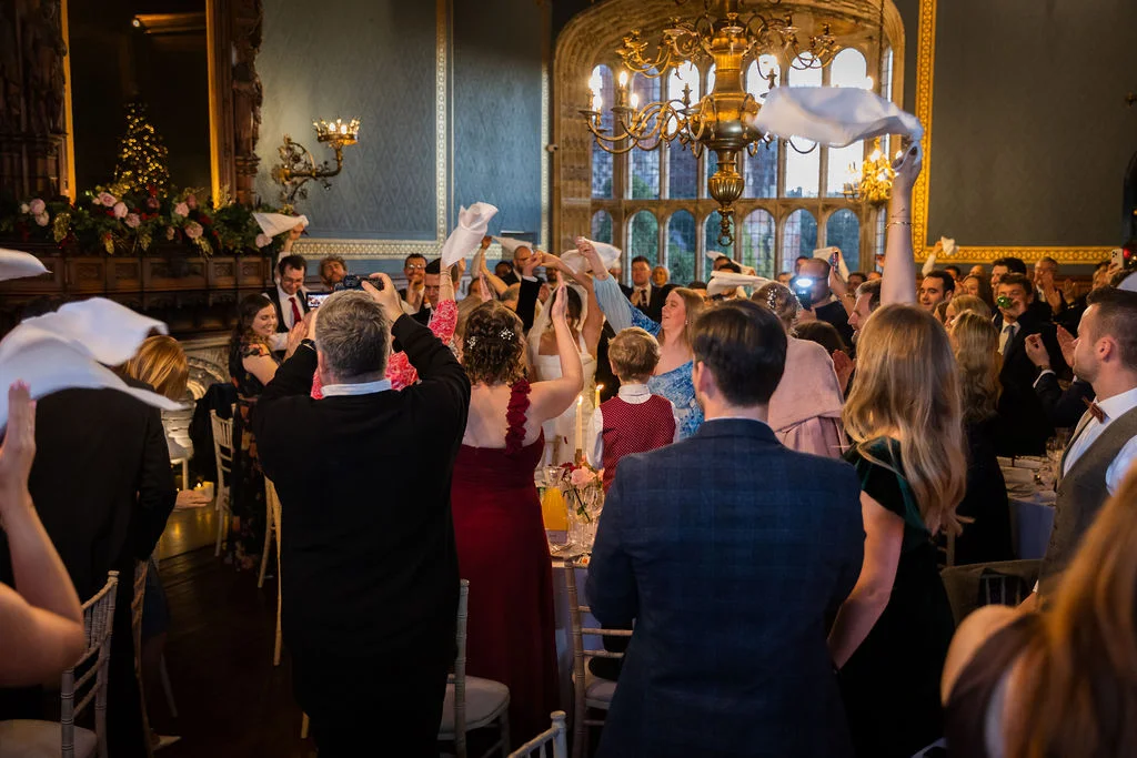 Wedding guests enthusiastically wave white napkins in the air during the wedding reception inside a grand, wood-panelled hall with a large ornate chandelier.