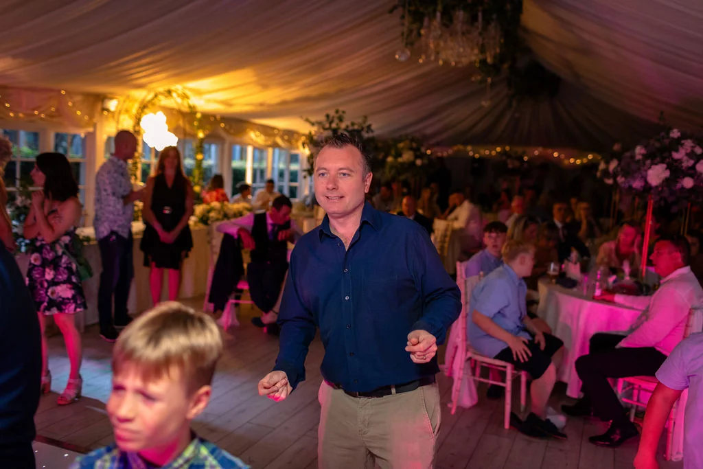 A wedding guest enjoys dancing on the illuminated dance floor inside a beautifully draped marquee reception filled with seated guests and warm lighting.