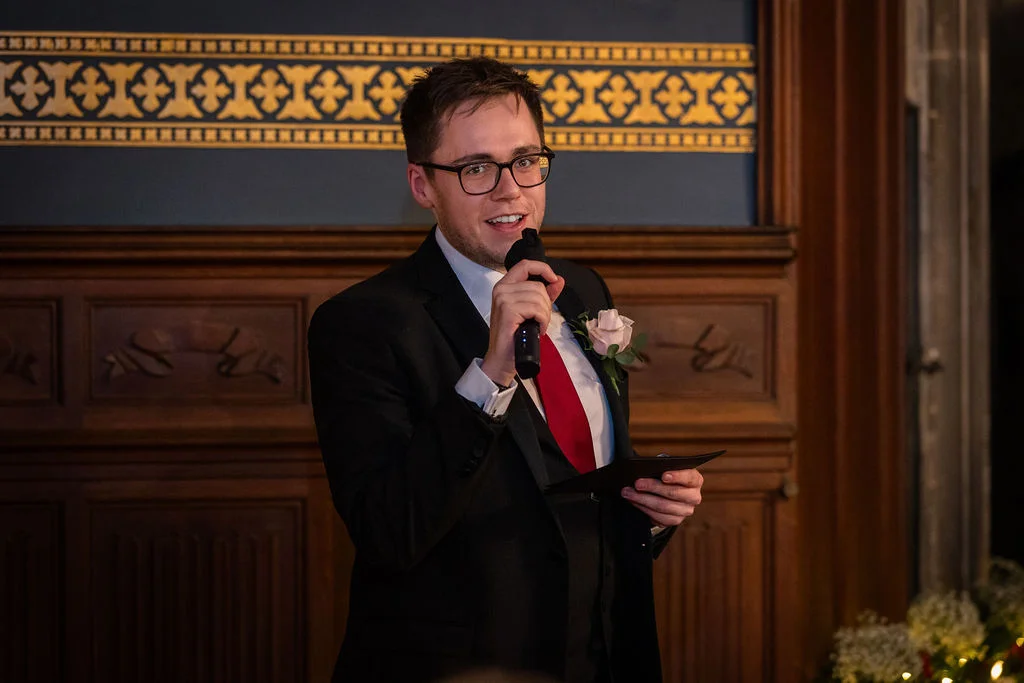 A groom wearing a three-piece black suit and a red tie delivers a speech into a microphone in a wood-panelled room.