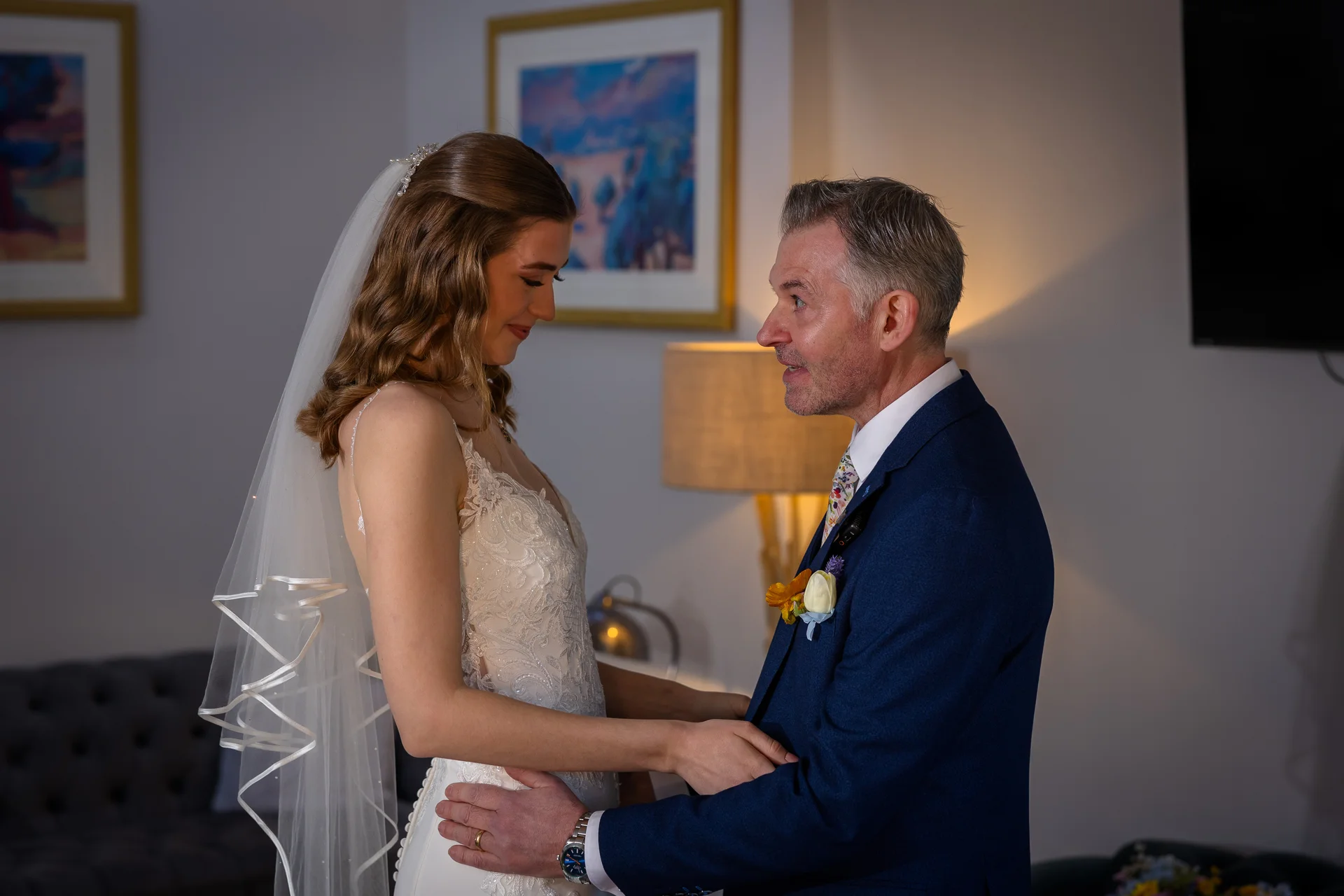 An emotional father of the bride shares a tender moment looking at his daughter in her lace-detailed gown and veil during morning preparations.