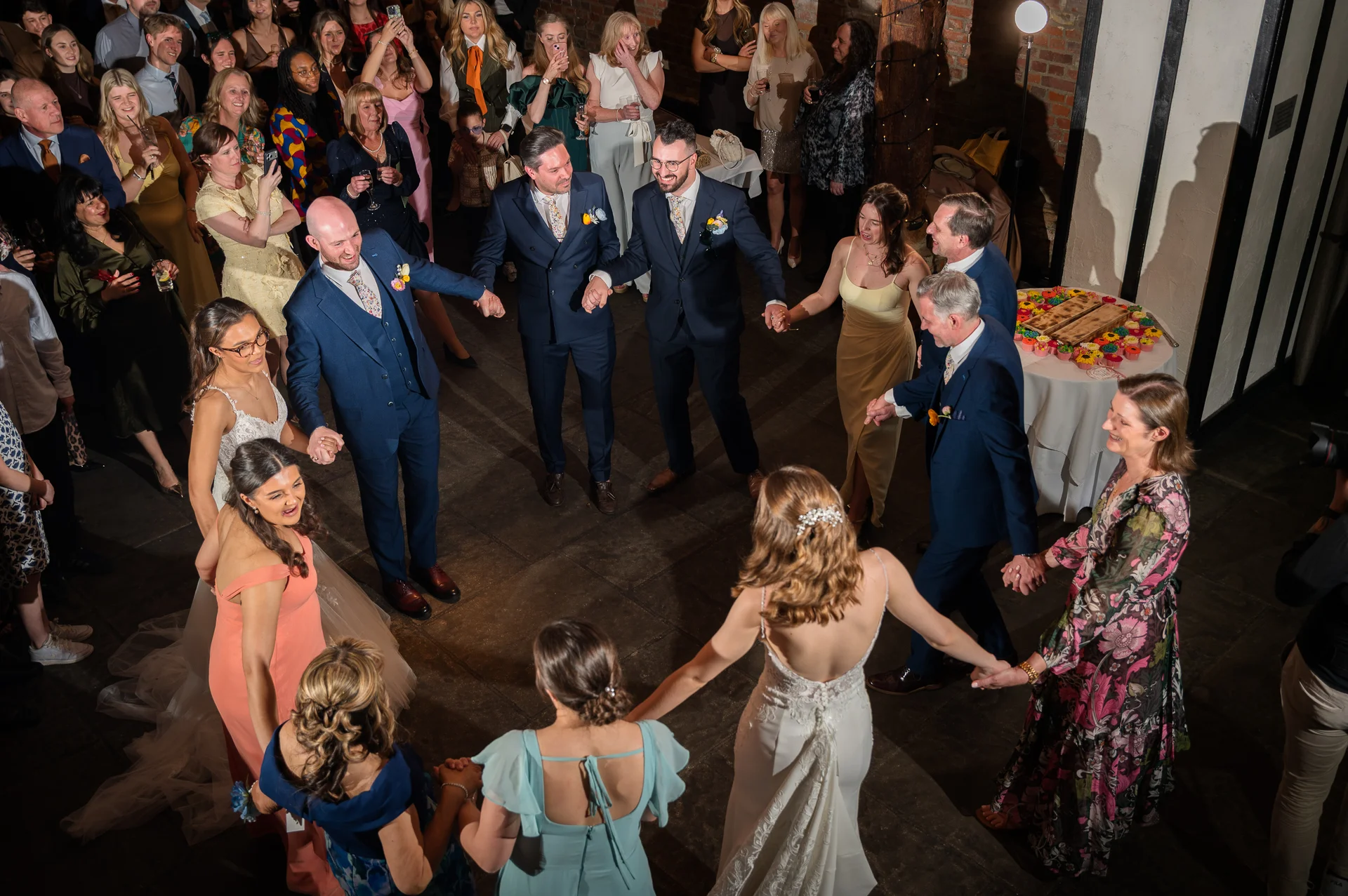 A high-angle shot captures a joyful circle of wedding guests and family holding hands and dancing together during an evening reception.