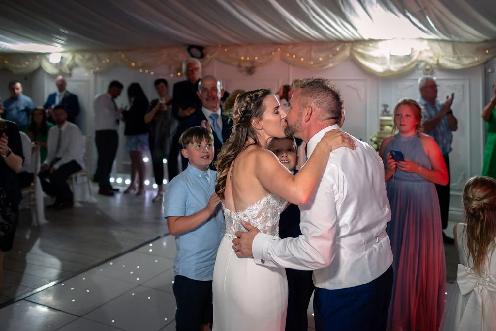 A newlywed couple shares a romantic kiss on an illuminated dance floor during their evening reception inside a draped marquee.