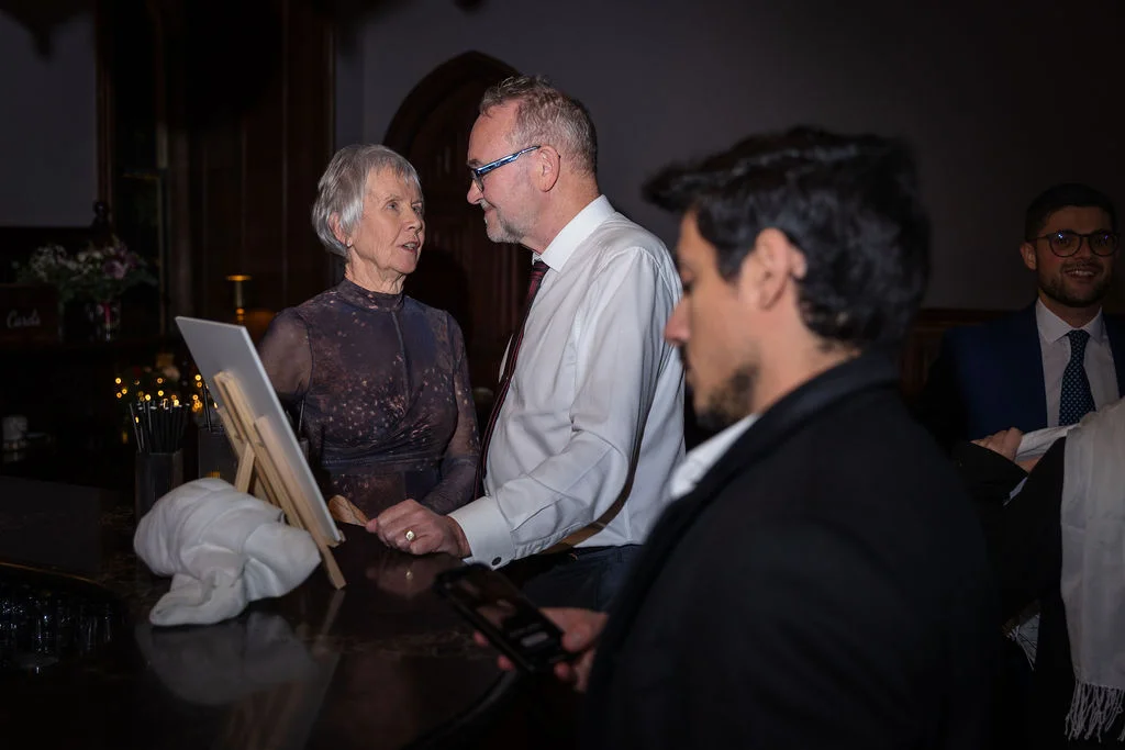 An older couple conversing across a bar area during a wedding evening reception, with other guests visible in the foreground and background.