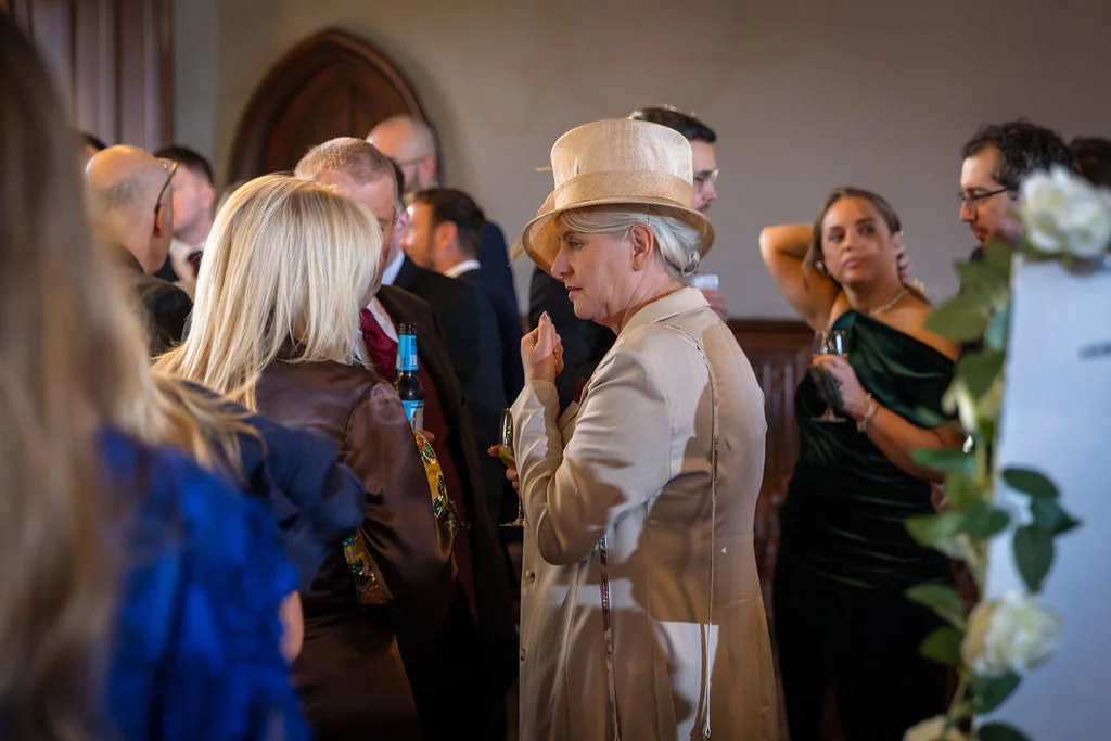 A distinguished female guest wearing a chic cream coat and matching hat socialises with other wedding attendees in a wood-panelled room.