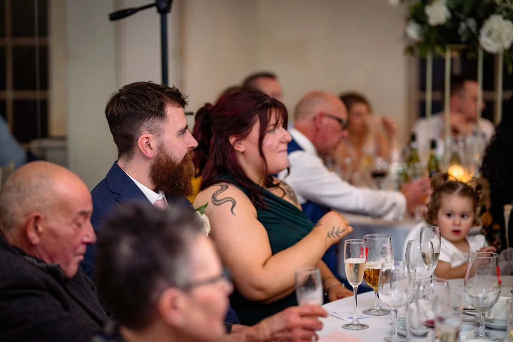 Wedding guests smiling and listening attentively during the speeches at a reception dinner table.