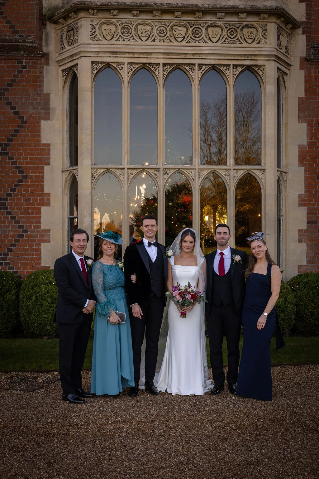 A bridal party poses formally outside the grand stone bay window of a historic country house venue.